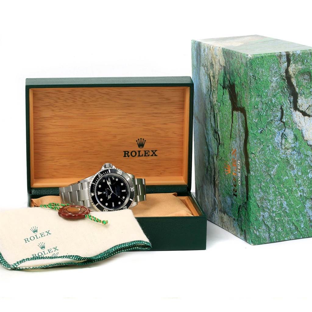 The image shows a Rolex Submariner 16610 Men
s Stainless Steel Black Dial 16610 Men
s Stainless Steel Black Dial watch in an open wooden box with a green cover and a cloth accessory.