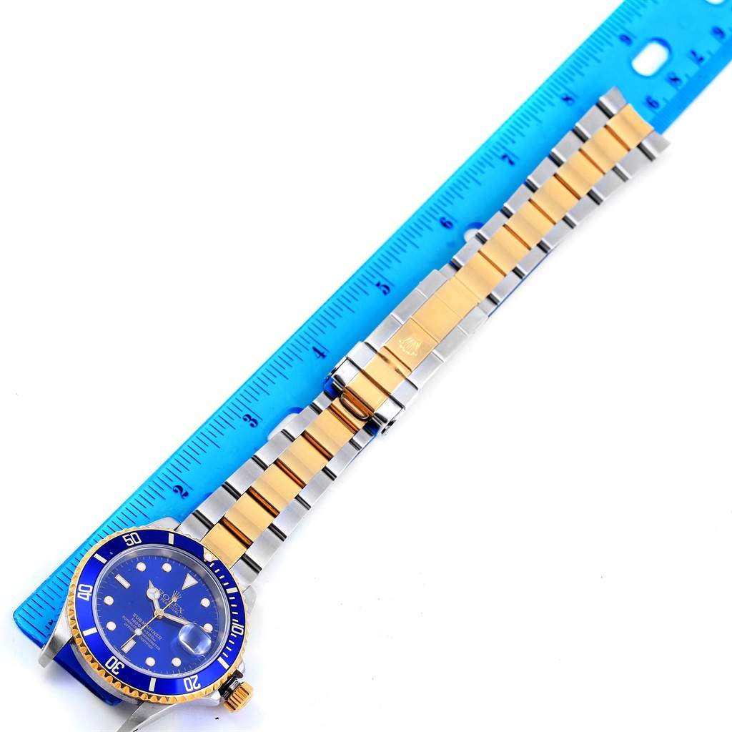 This image shows a Rolex Submariner 16613 Men
s Steel and Gold (two tone) Blue Dial 16613 Men
s Steel and Gold (two tone) Blue Dial watch laying flat next to a blue ruler, displaying the face, bezel, and two-tone bracelet.