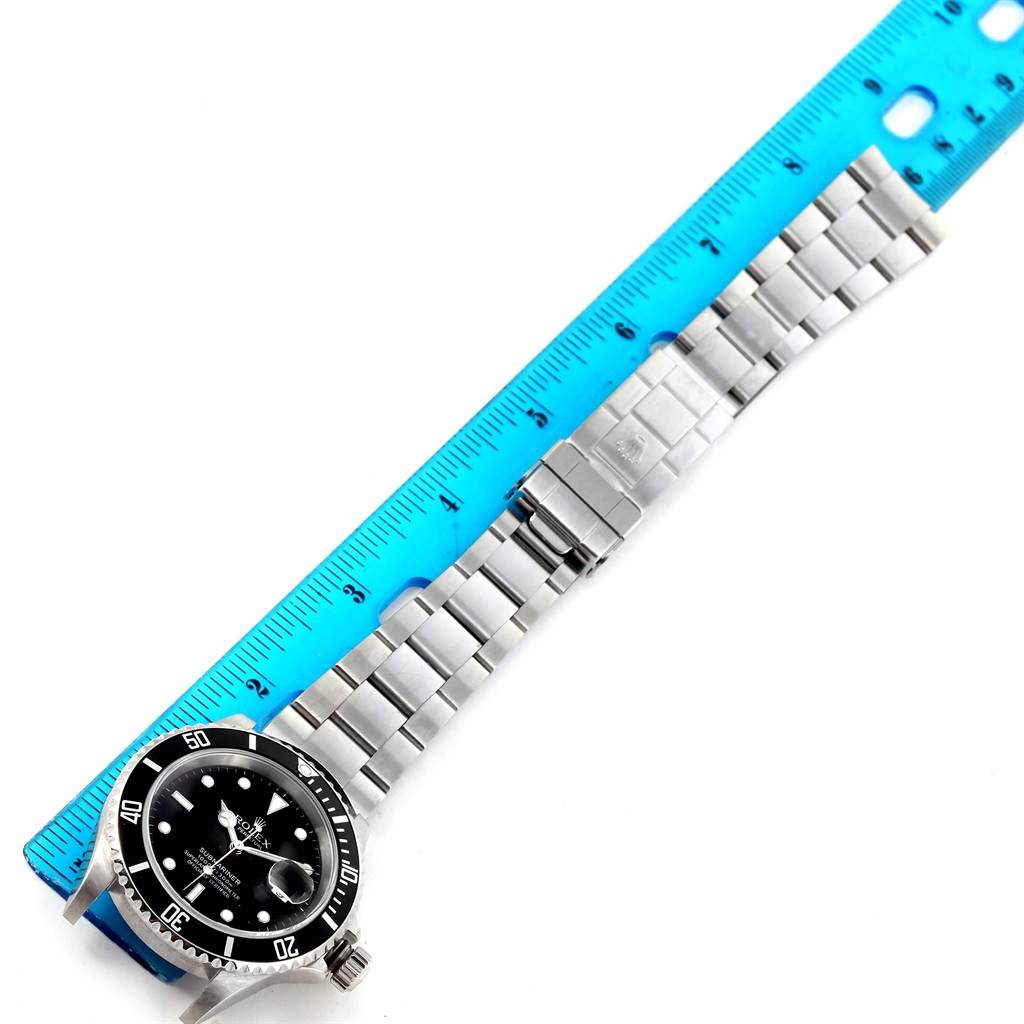 The image shows a Rolex Submariner 16610 Men
s Stainless Steel Black Dial 16610 Men
s Stainless Steel Black Dial watch face and stainless steel bracelet next to a blue ruler, laid flat.