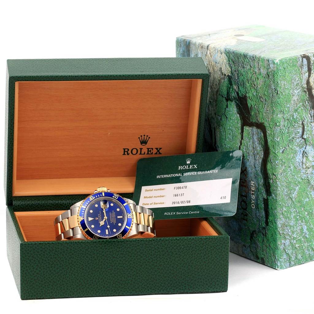 The image shows a Rolex Submariner 16613 Men's Steel and Gold (two tone) Blue Dial 16613 Men's Steel and Gold (two tone) Blue Dial watch placed in its open green box, along with its international service guarantee card.