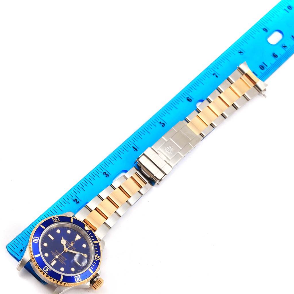 The Rolex Submariner 16613 Men's Steel and Gold (two tone) Blue Dial 16613 Men's Steel and Gold (two tone) Blue Dial watch is shown face-up with its full bracelet aligned next to a blue ruler for size reference.