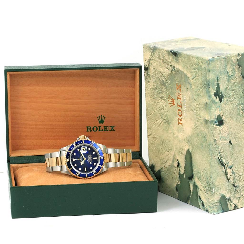 The image shows a Rolex Submariner 16613 Men's Steel and Gold (two tone) Blue Dial 16613 Men's Steel and Gold (two tone) Blue Dial watch placed inside an open green Rolex box with its outer packaging nearby.