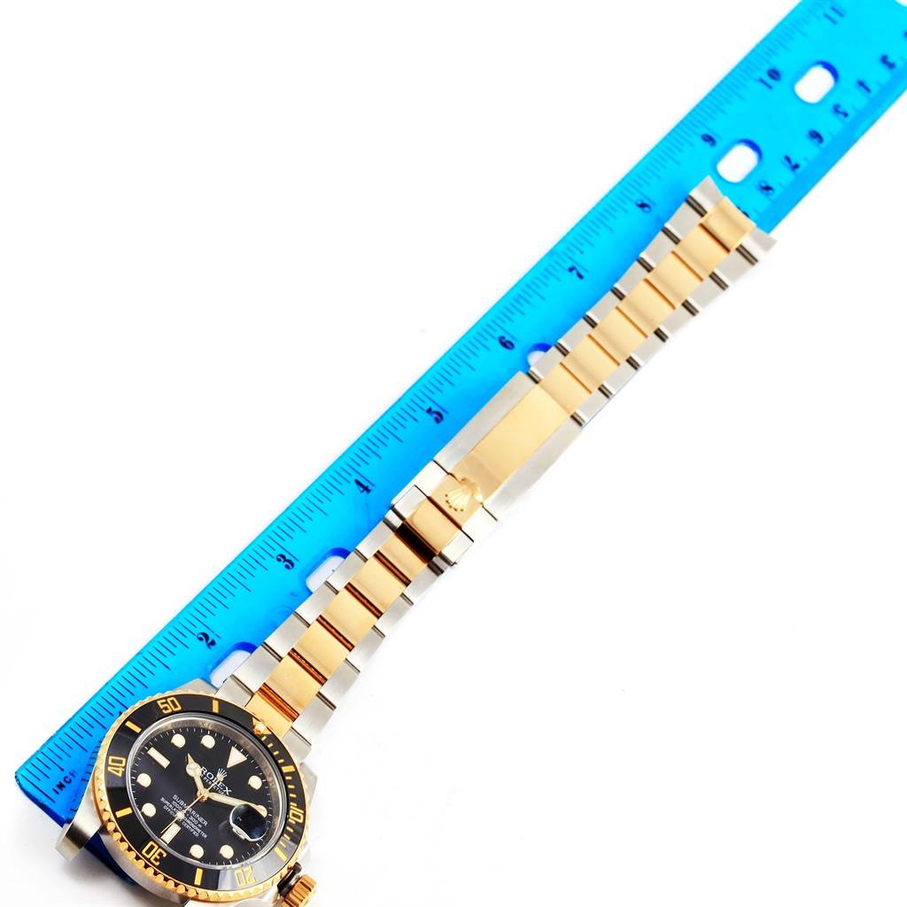 The image shows a Rolex Submariner 116613 Men
s Steel and Gold (two tone) Black Dial 116613 Men
s Steel and Gold (two tone) Black Dial watch laid flat alongside a blue ruler, highlighting its band and face.
