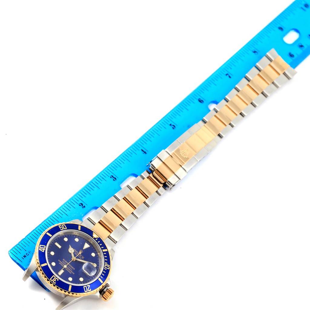 The Rolex Submariner 16613 Men
s Steel and Gold (two tone) Blue Dial 16613 Men
s Steel and Gold (two tone) Blue Dial watch is displayed flat, featuring its dial, bezel, and bracelet alongside a blue ruler.