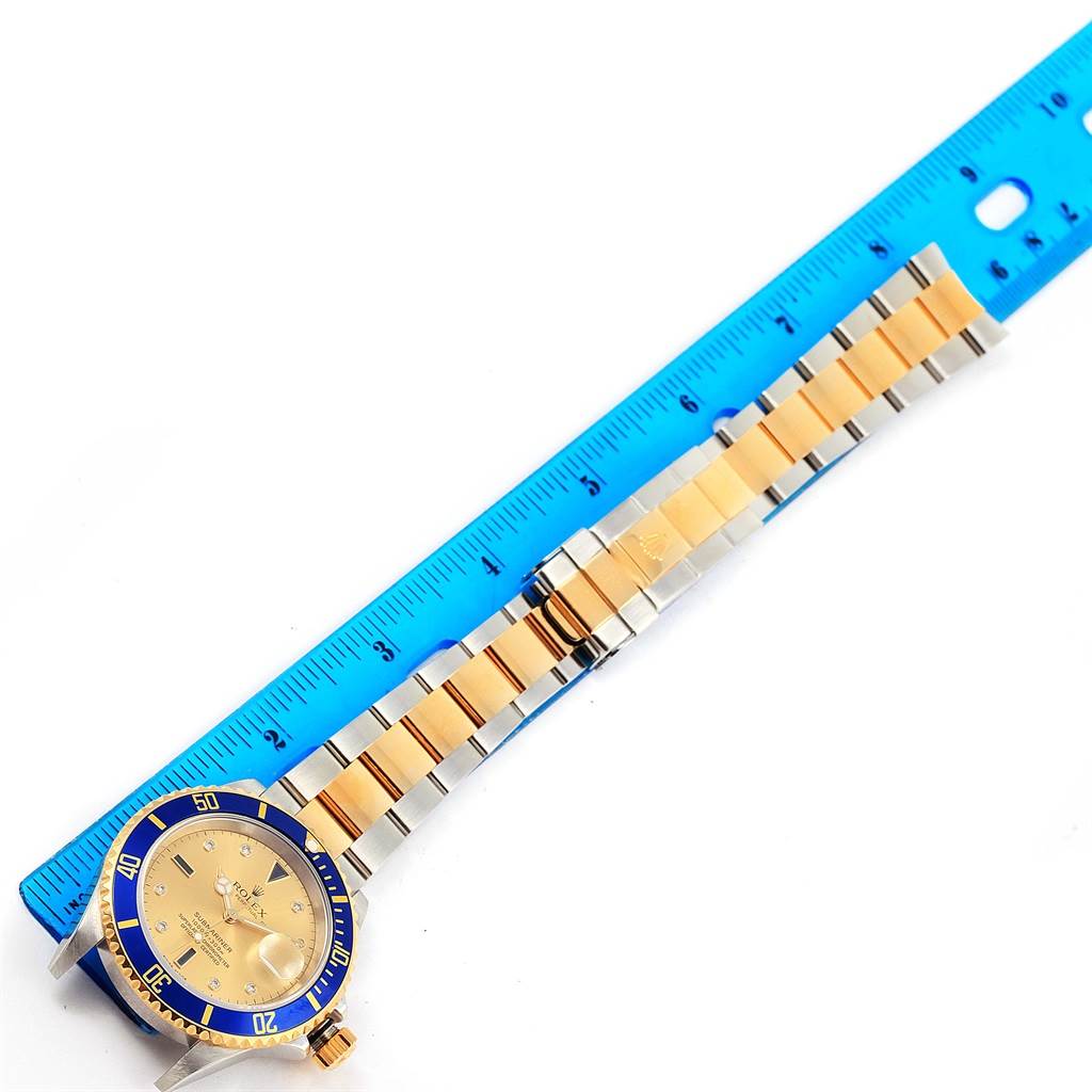 The image shows a Rolex Submariner 16613 Men
s Steel and Gold (two tone) Champagne Dial 16613 Men
s Steel and Gold (two tone) Champagne Dial watch laid flat, focusing on its face, bezel, and band next to a blue ruler for scale.