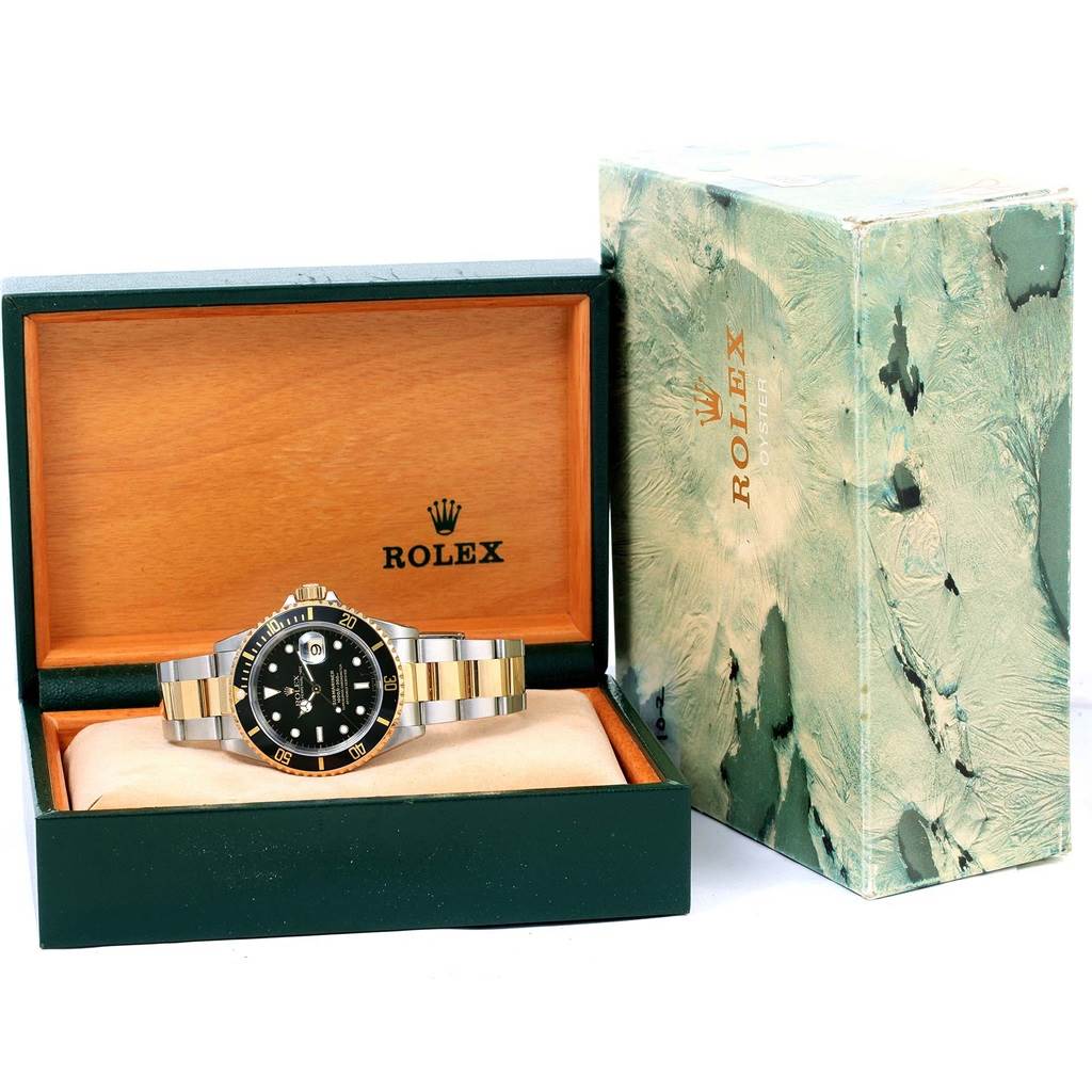 The Rolex Submariner 16613 Men
s Steel and Gold (two tone) Black Dial 16613 Men
s Steel and Gold (two tone) Black Dial watch is displayed front-on in its open wooden box with its packaging beside it.