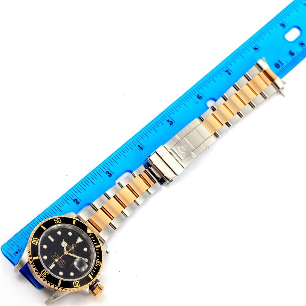 The image shows a Rolex Submariner 16613 Men
s Steel and Gold (two tone) Black Dial 16613 Men
s Steel and Gold (two tone) Black Dial watch laid flat next to a blue ruler, displaying its face and two-tone metal bracelet.