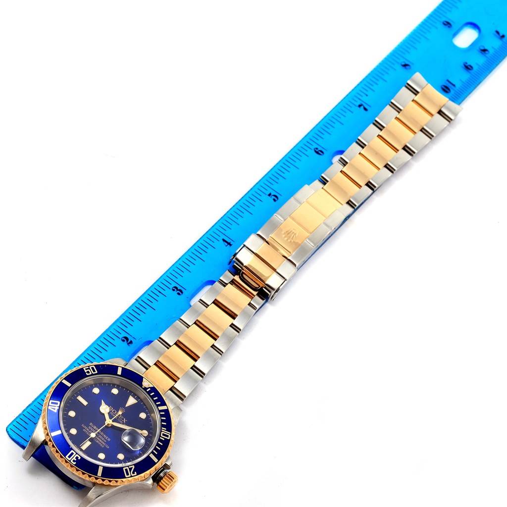 The image shows a Rolex Submariner 16613 Men's Steel and Gold (two tone) Blue Dial 16613 Men's Steel and Gold (two tone) Blue Dial watch laid flat next to a blue ruler, displaying the watch face and the metal bracelet.