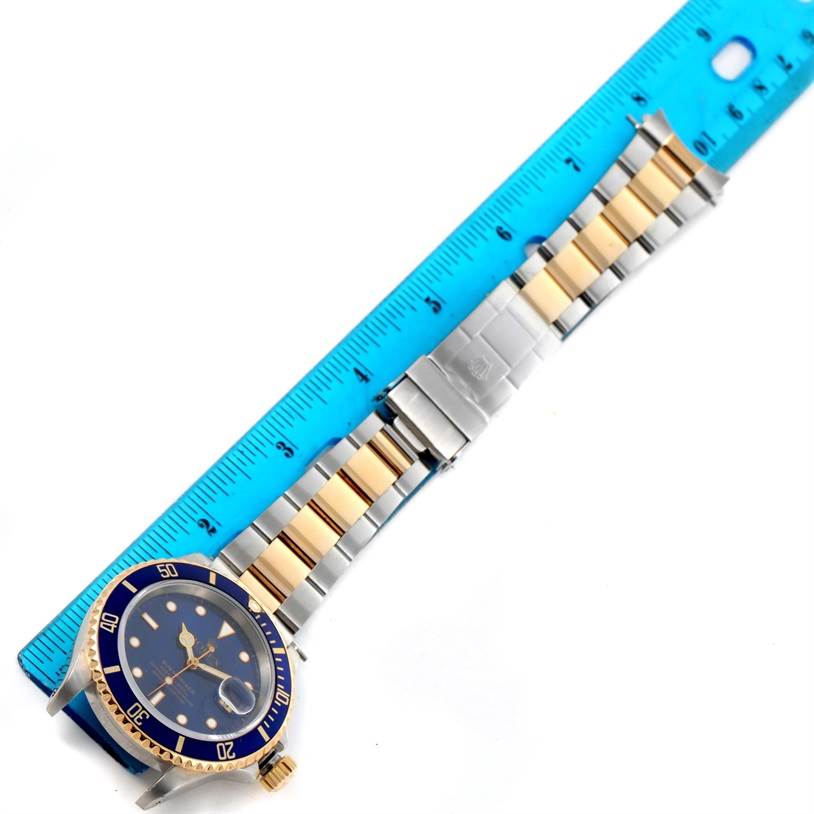 The image shows a top-down view of a Rolex Submariner 16613 Men
s Steel and Gold (two tone) Black Dial 16613 Men
s Steel and Gold (two tone) Black Dial watch with its blue dial, gold bezel, and two-tone bracelet measured against a blue ruler.