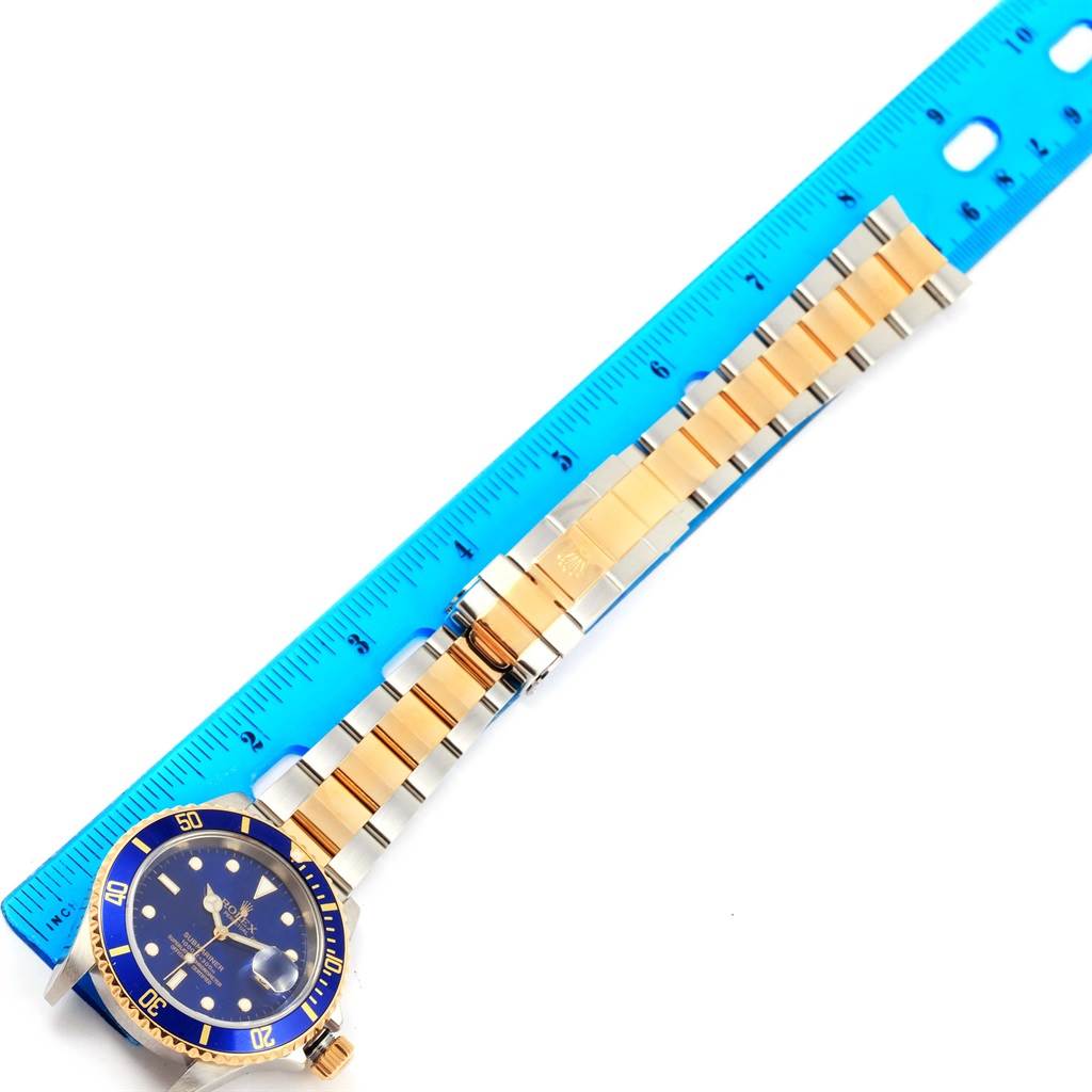 This image shows a Rolex Submariner 16613 Men's Steel and Gold (two tone) Blue Dial 16613 Men's Steel and Gold (two tone) Blue Dial watch laid flat next to a blue ruler, highlighting the gold and silver band and blue bezel.