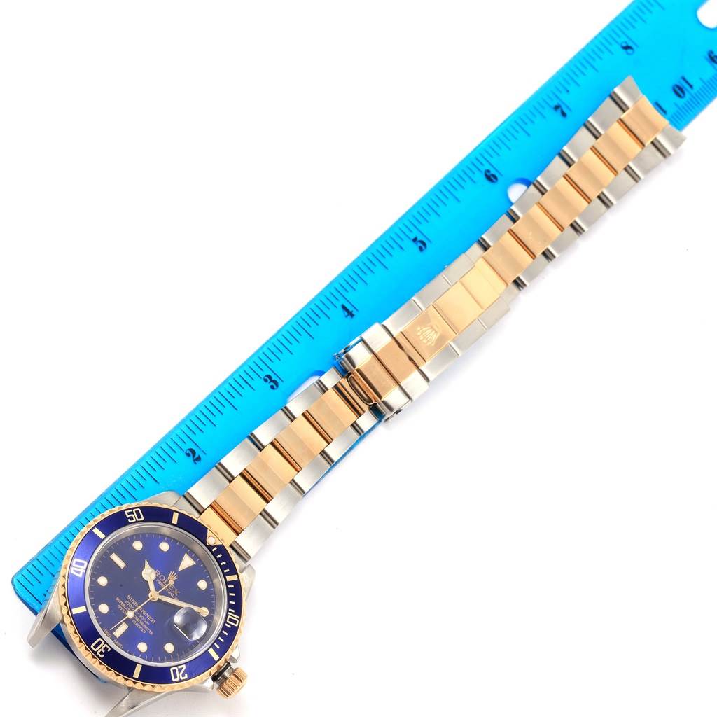 This image shows a Rolex Submariner 16613 Men's Steel and Gold (two tone) Blue Dial 16613 Men's Steel and Gold (two tone) Blue Dial watch with a blue dial and gold-tone bracelet, alongside a blue ruler.