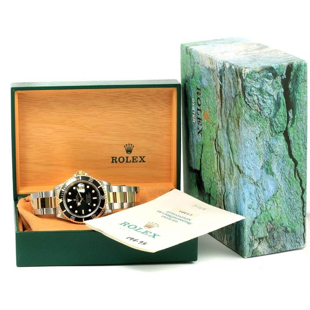 The image shows a Rolex Submariner 16613 Men
s Steel and Gold (two tone) Black Dial 16613 Men
s Steel and Gold (two tone) Black Dial watch in its open box along with accompanying documents and the outer green box.