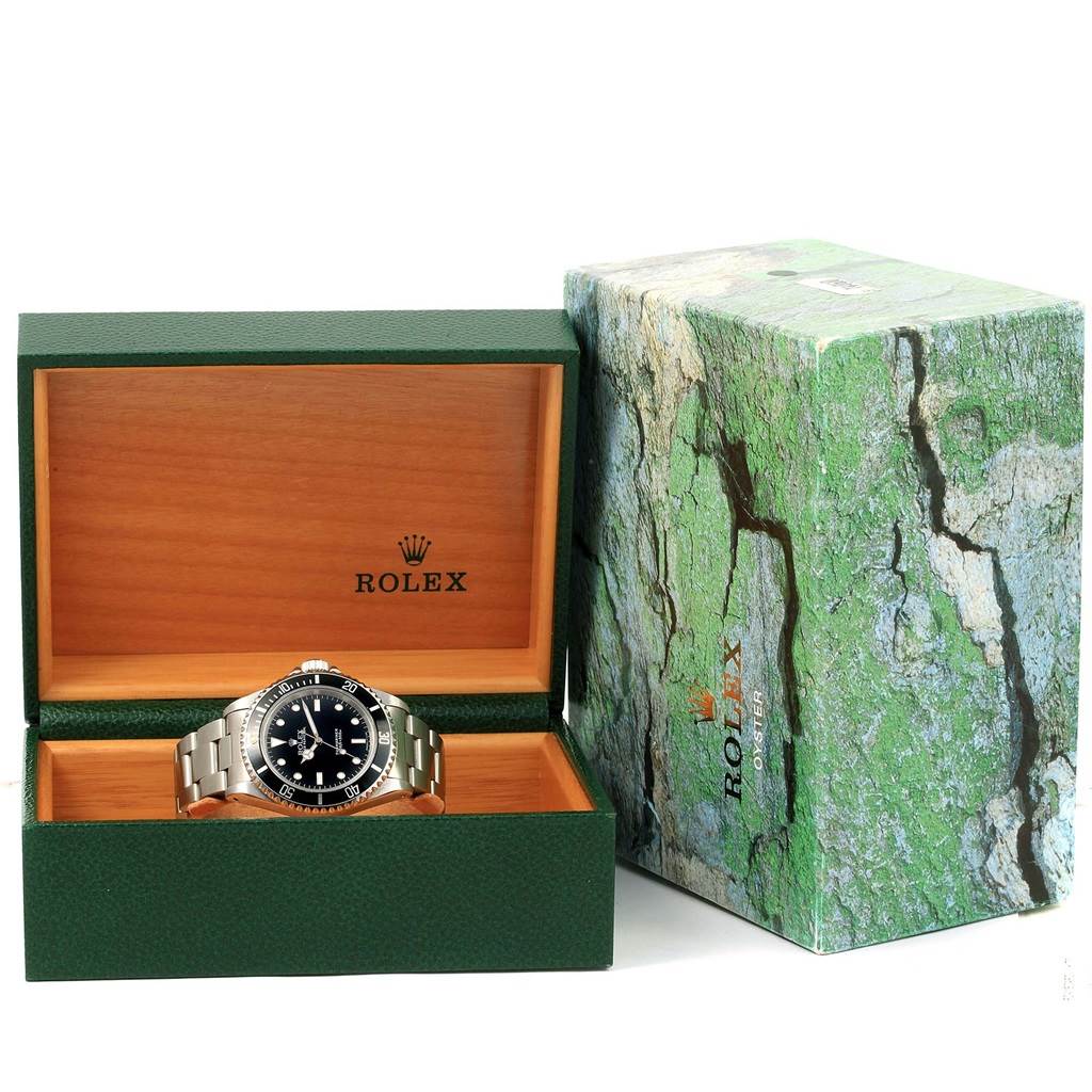The image shows a Rolex Submariner 14060 Men
s Stainless Steel Black Dial 14060 Men
s Stainless Steel Black Dial watch presented in an open green and wooden box, with its packaging next to it.