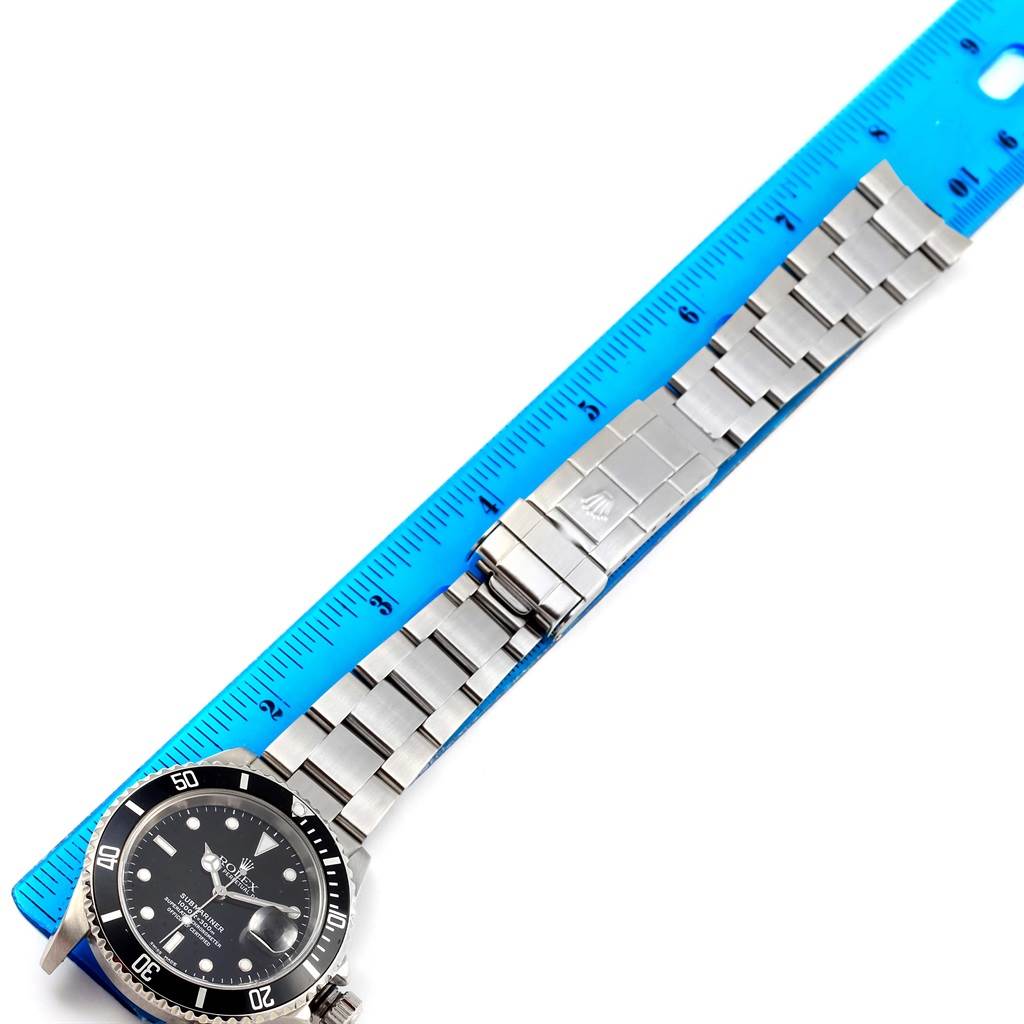 The image shows a Rolex Submariner 16610 Men's Stainless Steel Black Dial 16610 Men's Stainless Steel Black Dial laid flat against a blue ruler, displaying the watch face, bracelet, and clasp.