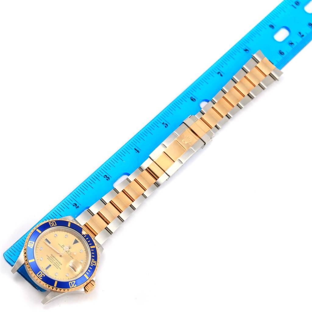 The Rolex Submariner 16613 Men's Steel and Gold (two tone) Champagne Dial 16613 Men's Steel and Gold (two tone) Champagne Dial watch is shown from a top-down angle, showcasing its face, bezel, and bracelet alongside a blue ruler.