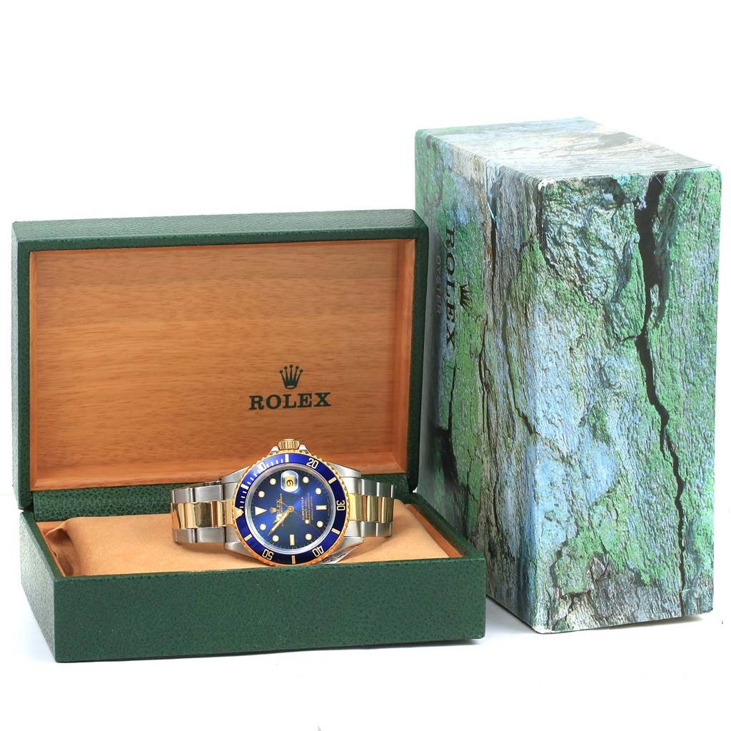 The Rolex Submariner 16613 Men
s Steel and Gold (two tone) Blue Dial 16613 Men
s Steel and Gold (two tone) Blue Dial watch is shown at a front angle, displayed in an open green and wood-finished box with its packaging visible.