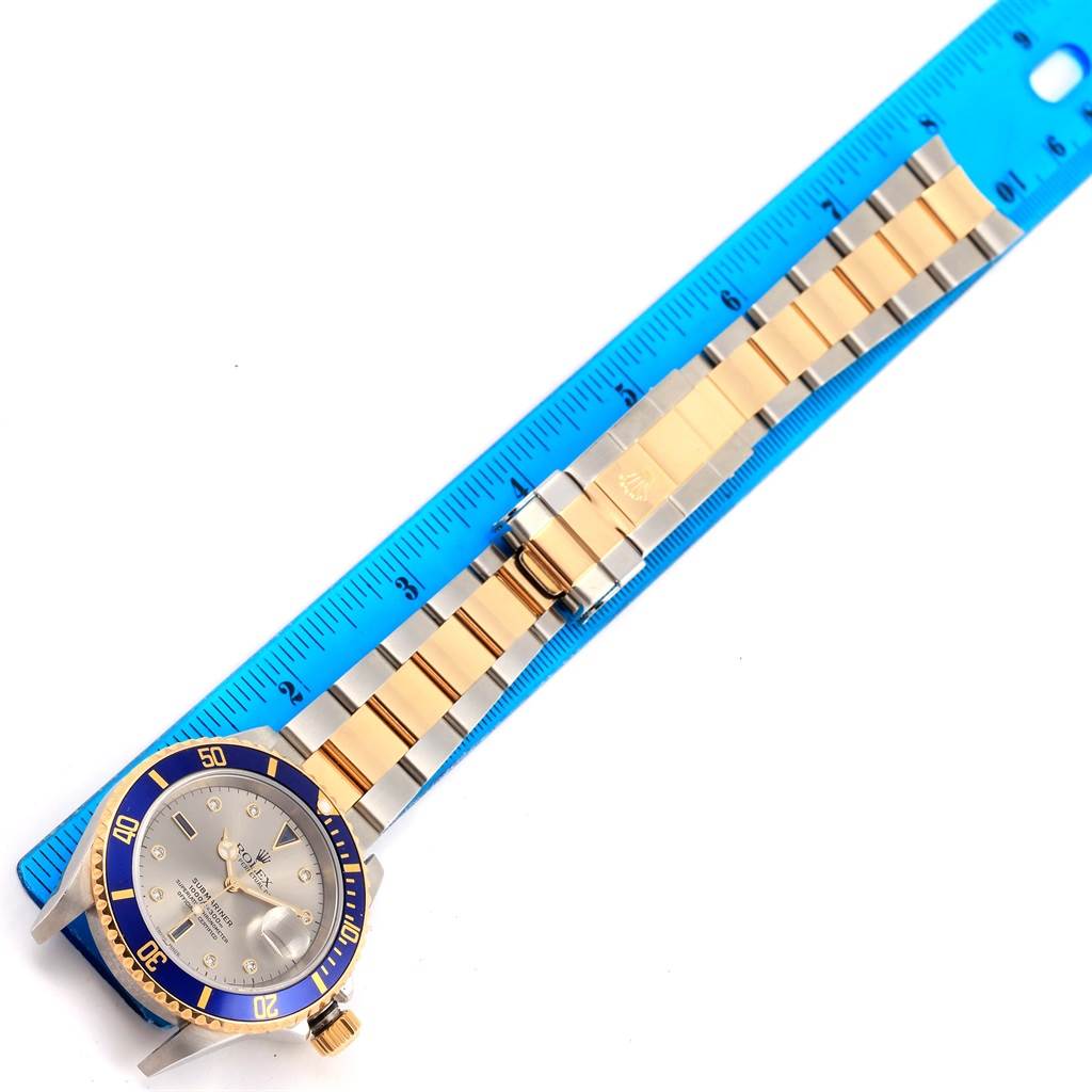 The image shows a Rolex Submariner 16613 Men
s Steel and Gold (two tone) Grey Dial 16613 Men
s Steel and Gold (two tone) Grey Dial watch laid flat with its band beside a blue ruler.
