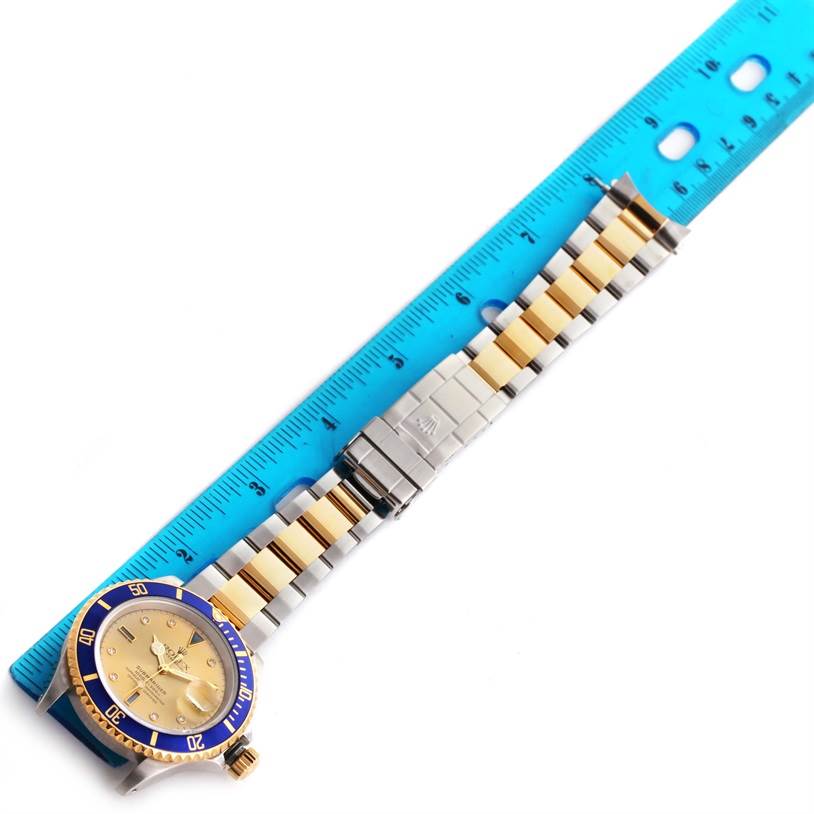 The image shows a Rolex Submariner 16613 Men's Steel and Gold (two tone) Champagne Dial 16613 Men's Steel and Gold (two tone) Champagne Dial watch laid flat next to a blue ruler, highlighting the two-tone bracelet and clasp.