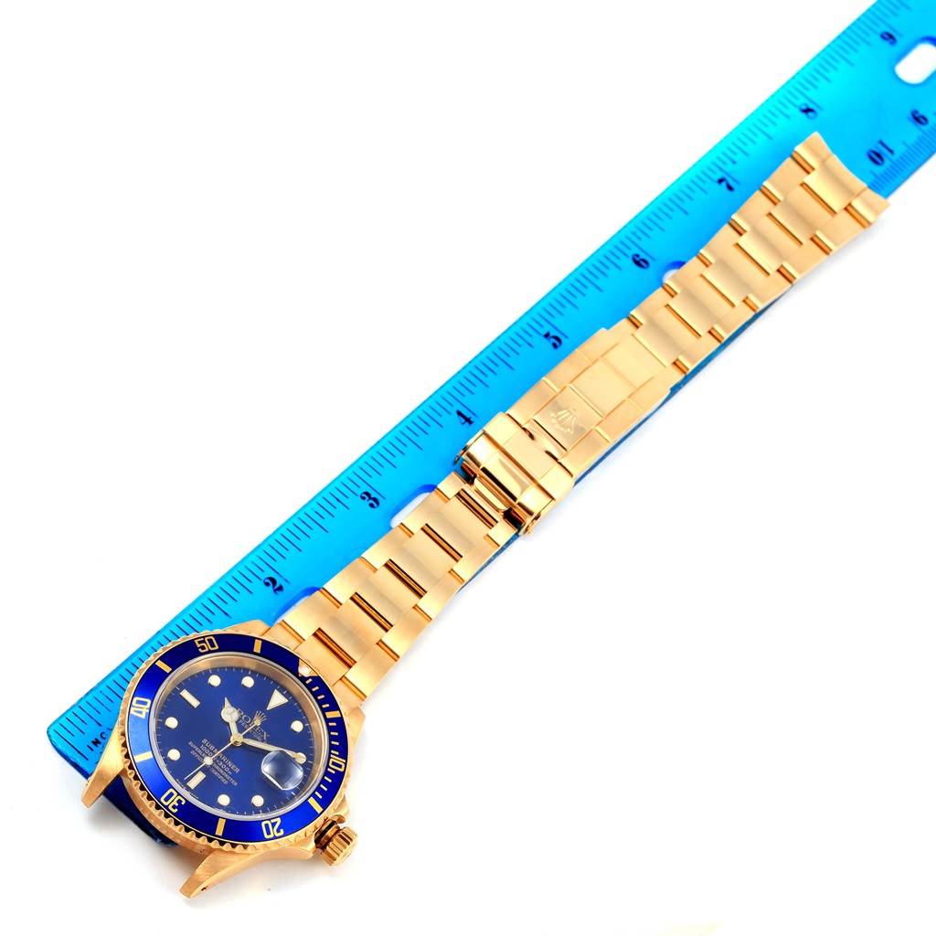 The Rolex Submariner 16618 Men's Yellow Gold Blue Dial 16618 Men's Yellow Gold Blue Dial watch is shown horizontally alongside a blue ruler, displaying its gold bracelet, clasp, and blue dial.
