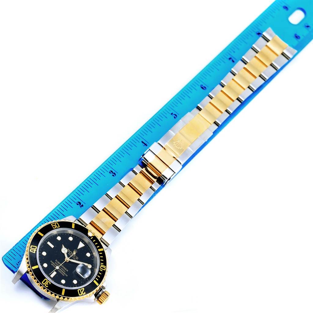 The image shows a top view of a Rolex Submariner 16613 Men's Stainless Steel Black Dial 16613 Men's Stainless Steel Black Dial watch with a gold and silver strap alongside a blue ruler for scale.
