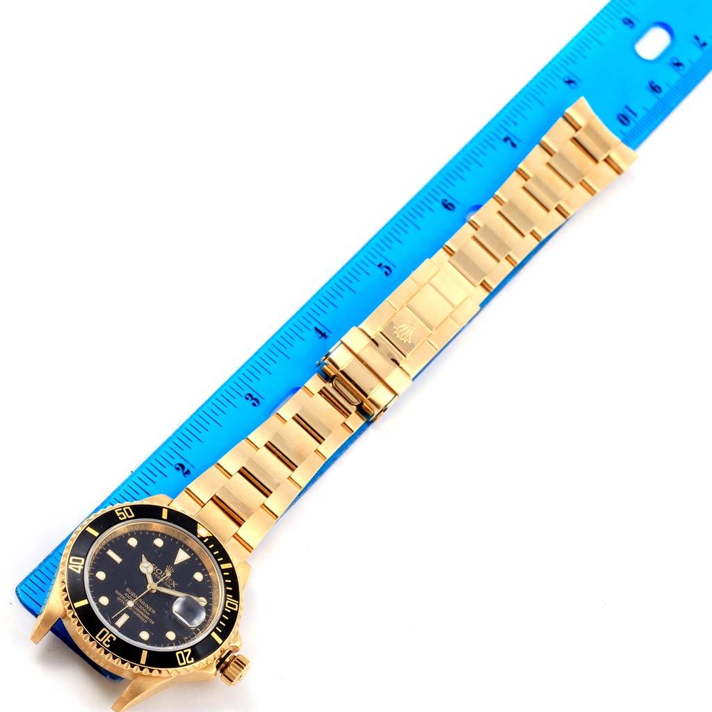The image shows a Rolex Submariner 16618 Men's Yellow Gold Black Dial 16618 Men's Yellow Gold Black Dial watch at an angle that highlights its gold bracelet, clasp, and black bezel, with a blue ruler for scale.