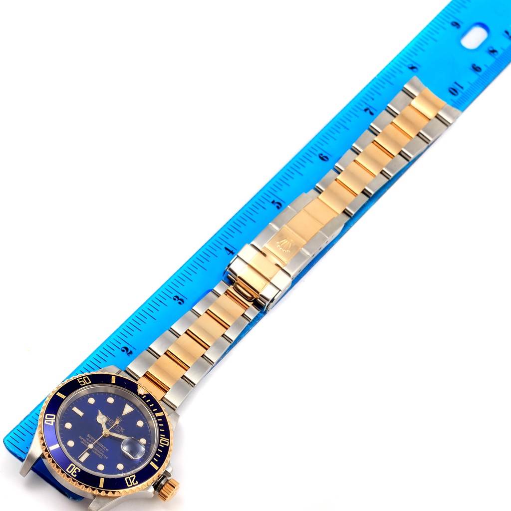 The image shows a Rolex Submariner 16613 Men's Steel and Gold (two tone) Blue Dial 16613 Men's Steel and Gold (two tone) Blue Dial watch laid flat with its bracelet extended next to a blue ruler for scale.