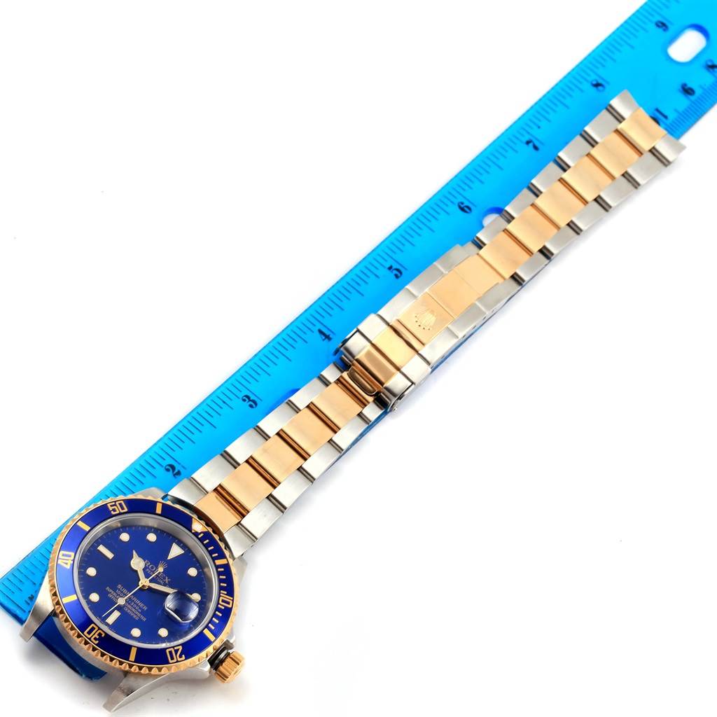 The image shows a Rolex Submariner 16613 Men's Steel and Gold (two tone) Blue Dial 16613 Men's Steel and Gold (two tone) Blue Dial watch laid flat with its band fully extended next to a blue ruler for measurement.