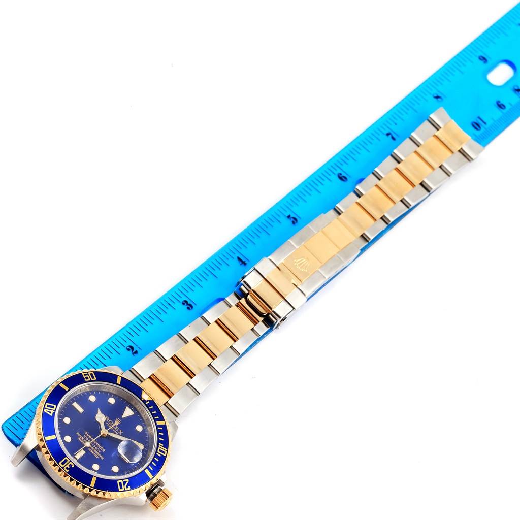 This image shows a Rolex Submariner 16613 Men's Steel and Gold (two tone) Blue Dial 16613 Men's Steel and Gold (two tone) Blue Dial at an overhead angle with its blue dial, bezel, and gold and silver bracelet next to a blue ruler.