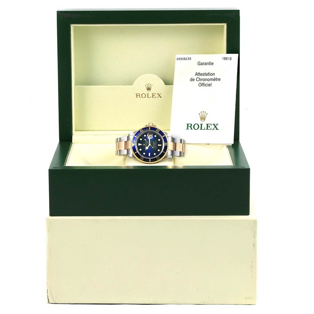 The image shows a Rolex Submariner 16613 Men's Steel and Gold (two tone) Blue Dial 16613 Men's Steel and Gold (two tone) Blue Dial watch in its open box, displaying the watch face, bracelet, and accompanying guarantee certificate.