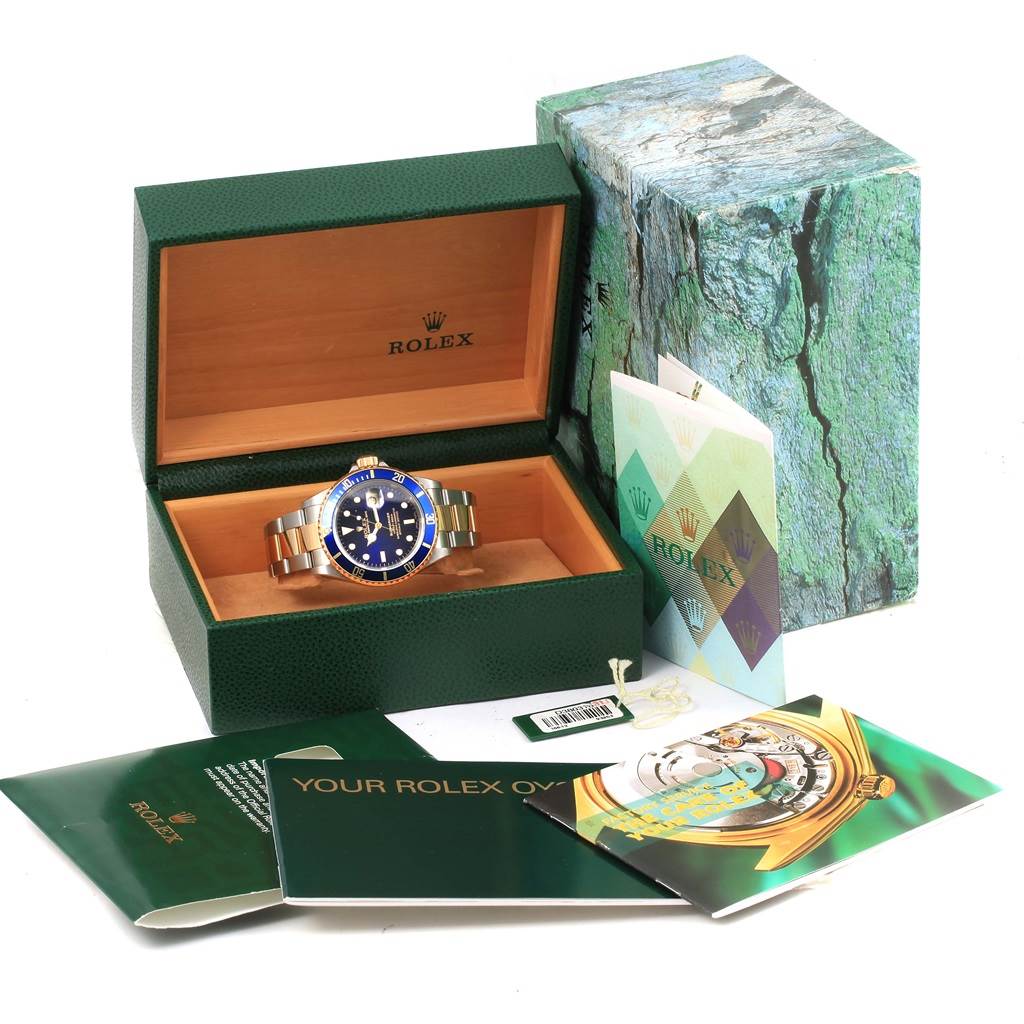 The image shows a Rolex Submariner 16613 Men's Steel and Gold (two tone) Blue Dial 16613 Men's Steel and Gold (two tone) Blue Dial watch at a slight angle in its open green box, accompanied by accessories like booklets and tags.