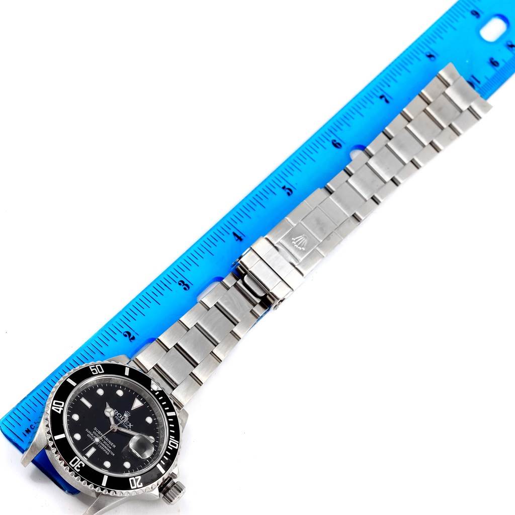 The Rolex Submariner 16610 Men's Stainless Steel Black Dial 16610 Men's Stainless Steel Black Dial watch is displayed flat with its stainless steel bracelet fully extended next to a blue ruler.