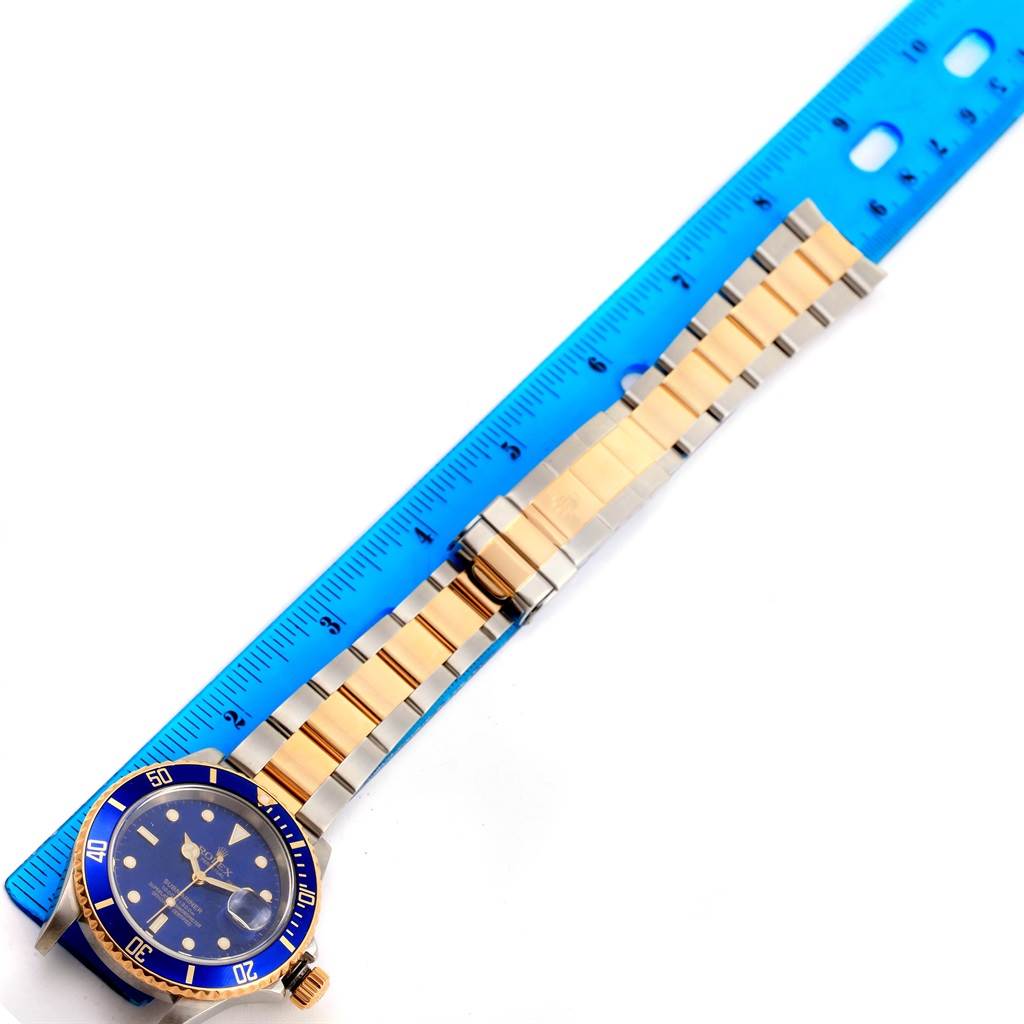 The Rolex Submariner 16613 Men
s Steel and Gold (two tone) Blue Dial 16613 Men
s Steel and Gold (two tone) Blue Dial watch is shown flat, face up, next to a blue ruler, displaying its gold and silver bracelet and blue dial.