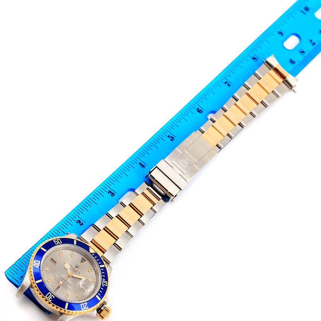 The Rolex Submariner 16613 Men's Steel and Gold (two tone) Grey Dial 16613 Men's Steel and Gold (two tone) Grey Dial watch is shown from a top-down angle, displaying its face, bezel, and bracelet against a blue ruler.