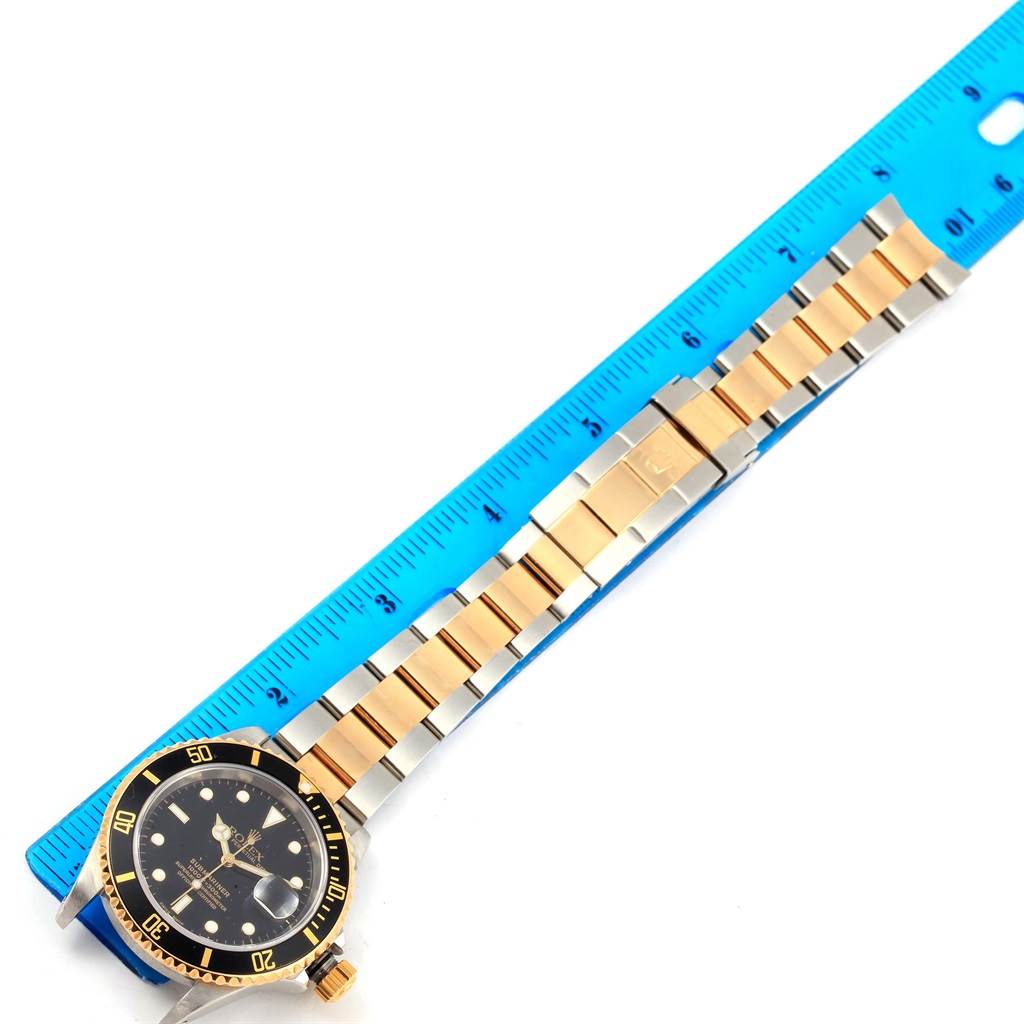 The image shows a Rolex Submariner 16613 Men
s Steel and Gold (two tone) Black Dial 16613 Men
s Steel and Gold (two tone) Black Dial watch with a gold and silver bracelet next to a blue ruler, viewing the front and side angle.