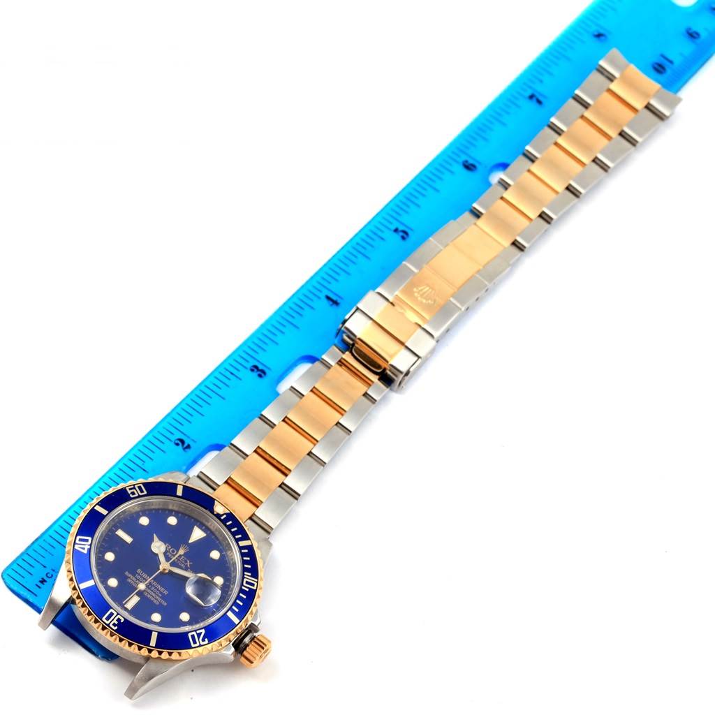 The image shows a Rolex Submariner 16613 Men's Steel and Gold (two tone) Blue Dial 16613 Men's Steel and Gold (two tone) Blue Dial watch laid flat with a blue dial and a two-tone bracelet next to a blue ruler.