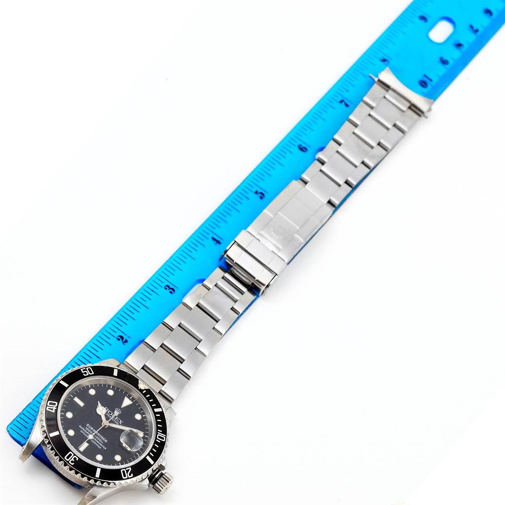 The image shows a Rolex Submariner 16610 Men's Stainless Steel Black Dial 16610 Men's Stainless Steel Black Dial watch, featuring the face and full length of the metal bracelet, with a blue ruler for scale.