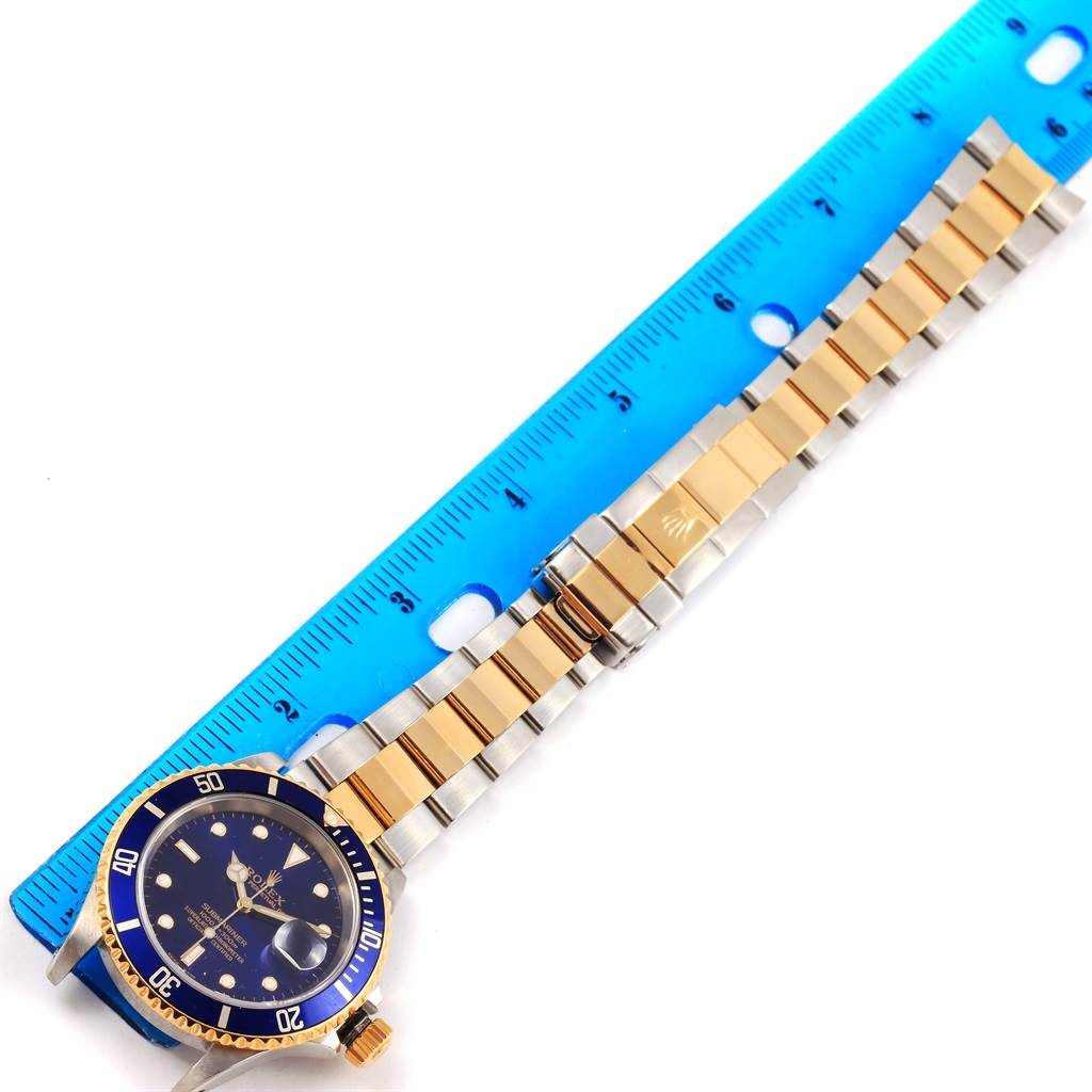 The image shows a Rolex Submariner 16613 Men's Steel and Gold (two tone) Blue Dial 16613 Men's Steel and Gold (two tone) Blue Dial watch laid flat with its face, bezel, crown, and full bracelet visible next to a blue ruler.