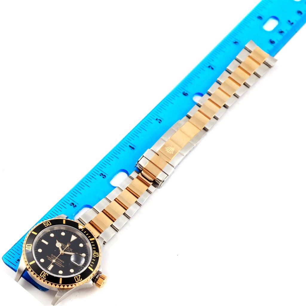 The image shows a Rolex Submariner 16613 Men's Steel and Gold (two tone) Black Dial 16613 Men's Steel and Gold (two tone) Black Dial watch, including the face, bezel, and full bracelet laid beside a blue ruler for scale.
