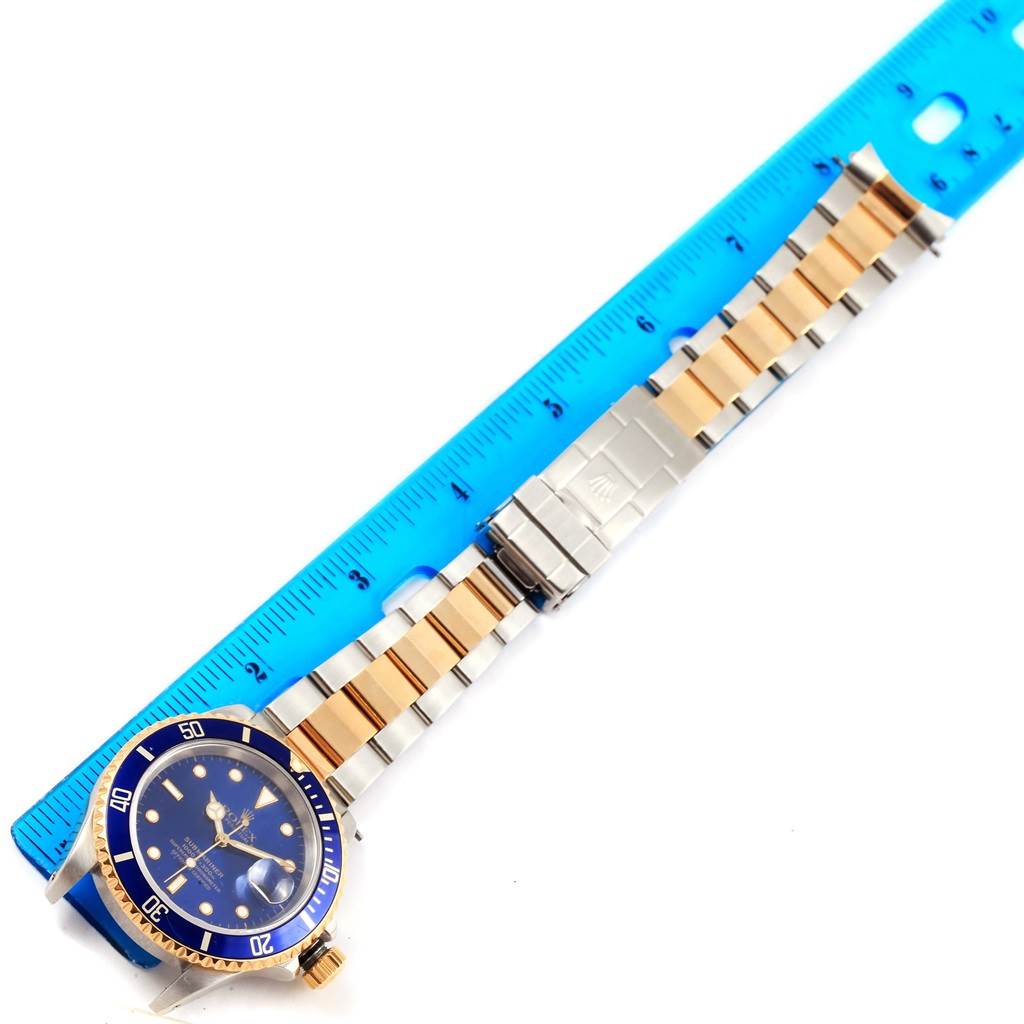 The image shows a Rolex Submariner 16613 Men's Steel and Gold (two tone) Blue Dial 16613 Men's Steel and Gold (two tone) Blue Dial watch with a blue dial and bezel, alongside a blue measure tape.