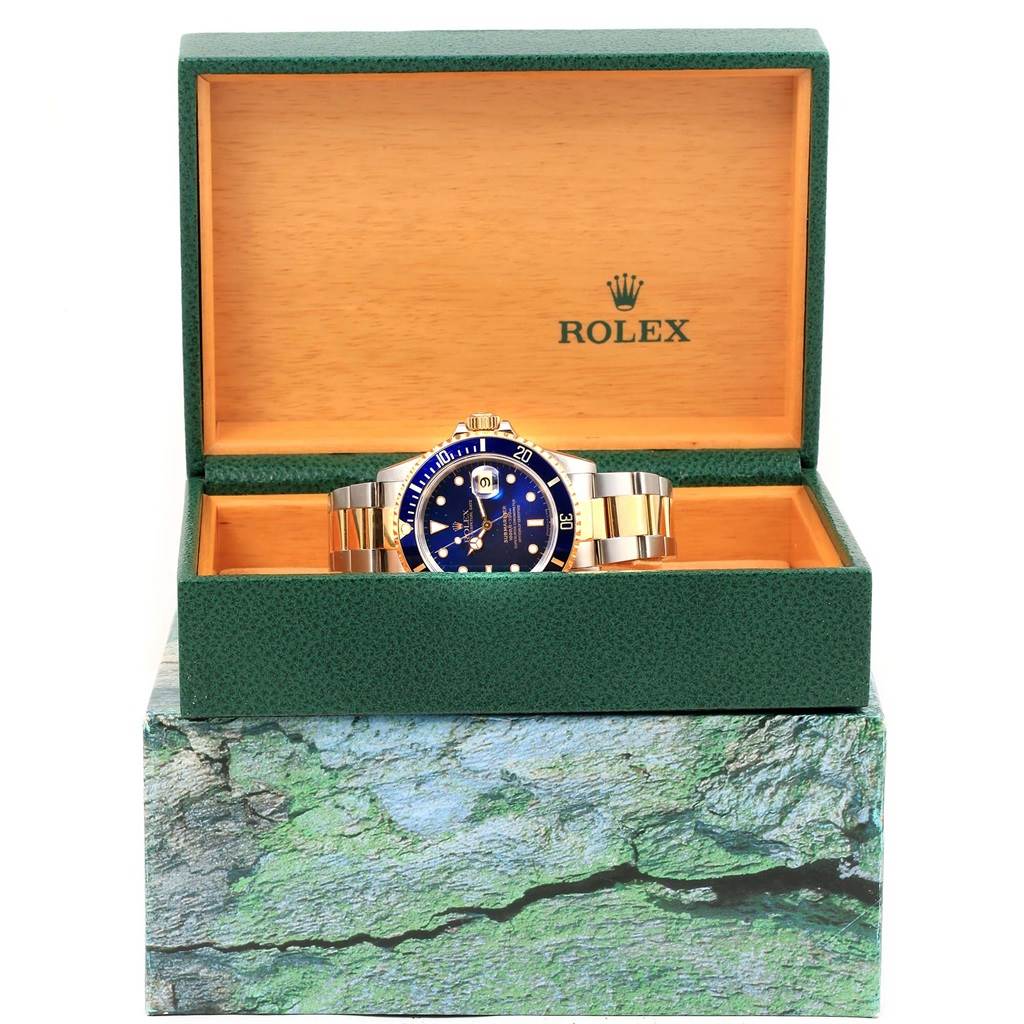 The Rolex Submariner 16613 Men's Steel and Gold (two tone) Blue Dial 16613 Men's Steel and Gold (two tone) Blue Dial watch is shown from the front inside its green and brown branded case.