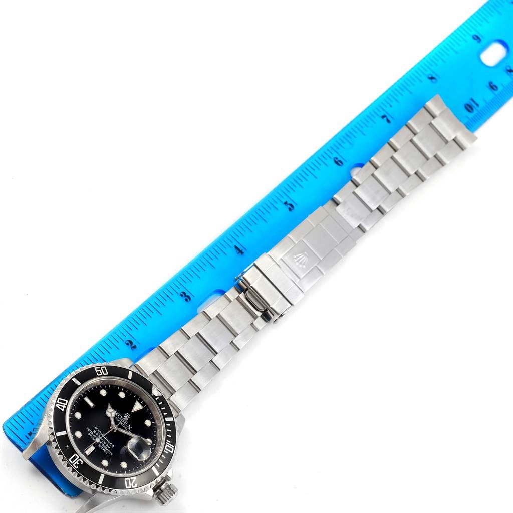 The image shows the Rolex Submariner 16610 Men
s Stainless Steel Black Dial 16610 Men
s Stainless Steel Black Dial watch at a top-down angle, along with a stainless steel bracelet and part of a blue ruler.