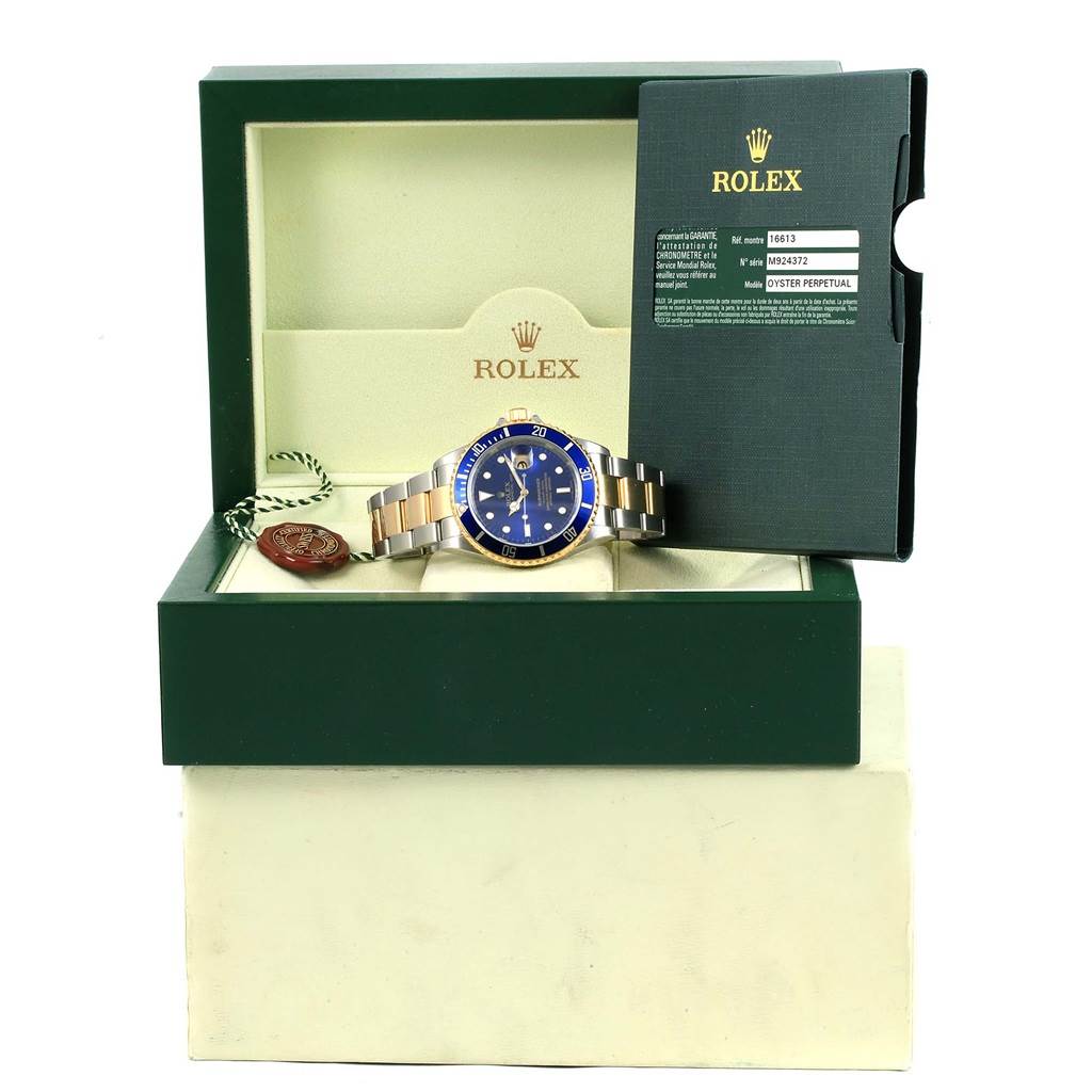This image shows a Rolex Submariner 16613 Men's Steel and Gold (two tone) Blue Dial 16613 Men's Steel and Gold (two tone) Blue Dial watch in its box with a booklet and hang tag from an angled front view.