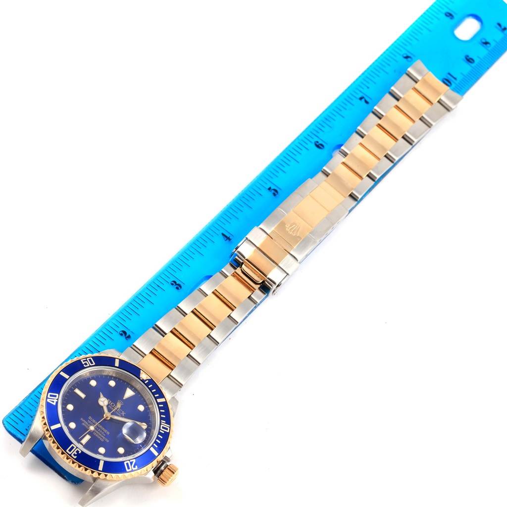 The image displays a top-down view of a Rolex Submariner 16613 Men's Steel and Gold (two tone) Blue Dial 16613 Men's Steel and Gold (two tone) Blue Dial watch with a blue dial and a two-tone bracelet next to a blue ruler.
