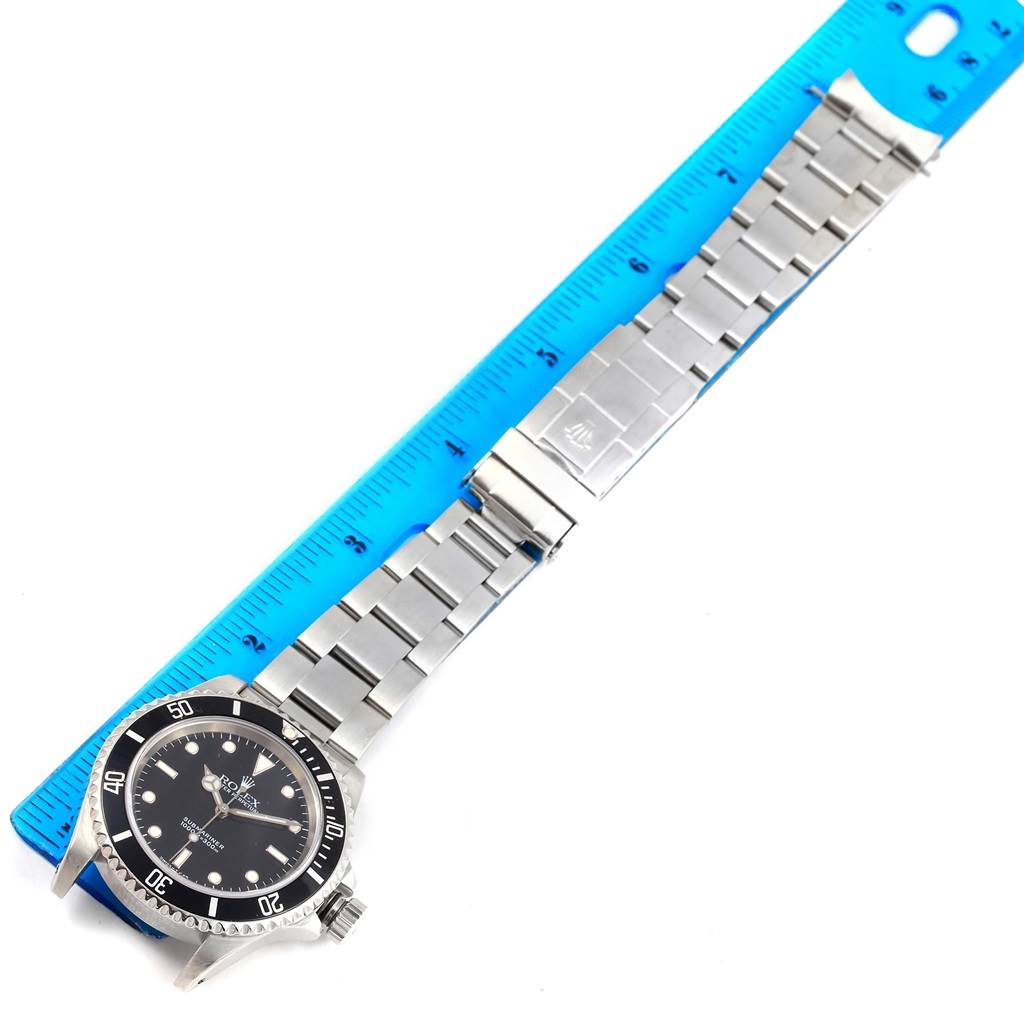 The angle shows the Rolex Submariner 14060 Men's Stainless Steel Black Dial 14060 Men's Stainless Steel Black Dial watch face and bracelet lying next to a blue ruler.