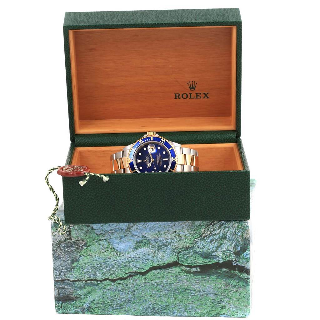 The Rolex Submariner 16613 Men
s Steel and Gold (two tone) Blue Dial 16613 Men
s Steel and Gold (two tone) Blue Dial watch is displayed in an open, green and wooden box, showing the face and bracelet prominently from a front angle.