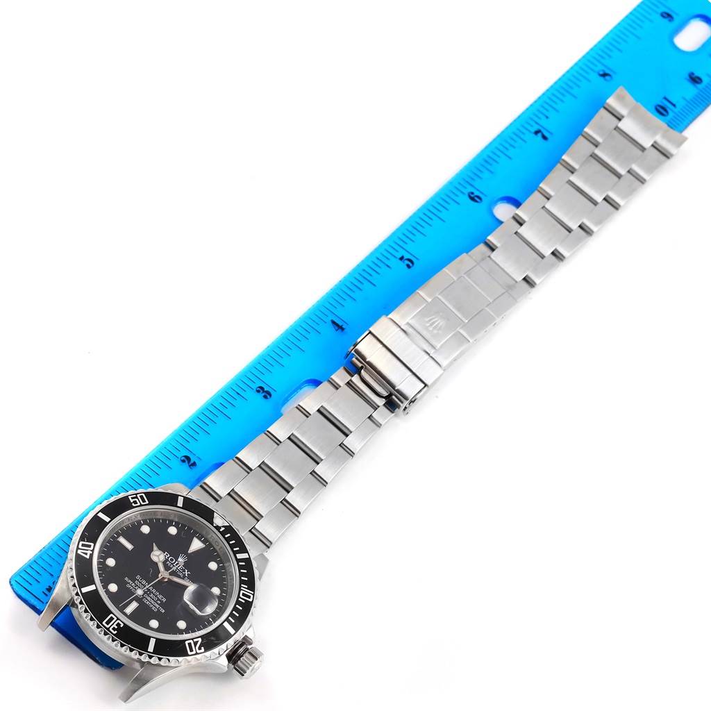 The Rolex Submariner 16610 Men's Stainless Steel Black Dial 16610 Men's Stainless Steel Black Dial watch is shown at an oblique angle, displaying the face, bezel, crown, and metal bracelet near a blue ruler.