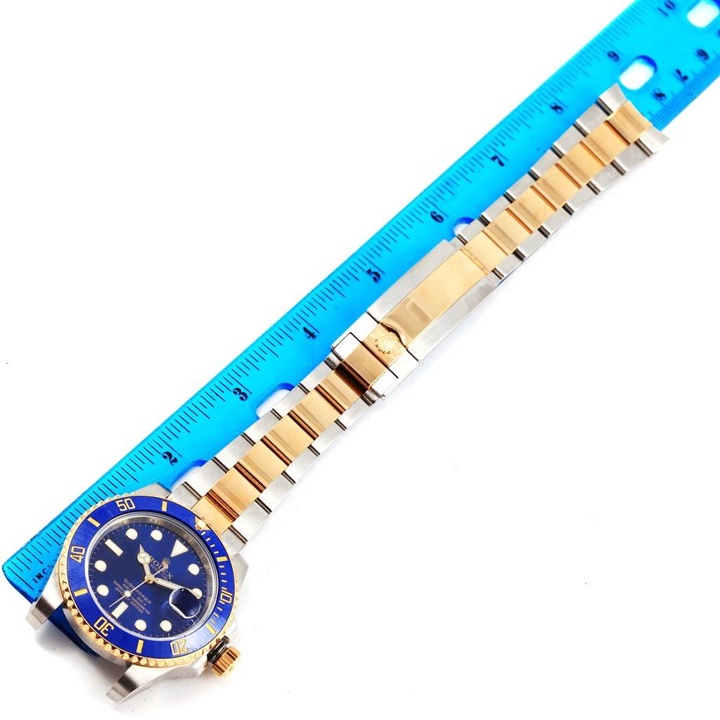 A Rolex Submariner 116613 Men's Steel and Gold (two tone) Blue Dial 116613 Men's Steel and Gold (two tone) Blue Dial watch is shown from a top-down angle alongside a blue ruler, displaying the case, dial, bezel, and bracelet.