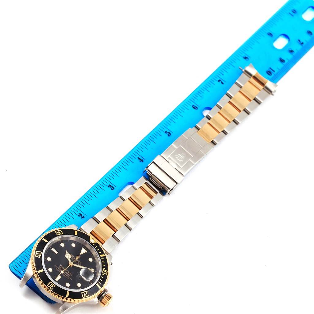 The image shows a Rolex Submariner 16613 Men
s Steel and Gold (two tone) Black Dial 16613 Men
s Steel and Gold (two tone) Black Dial from a top-down angle, showcasing the watch face, bezel, and two-tone bracelet alongside a blue ruler.