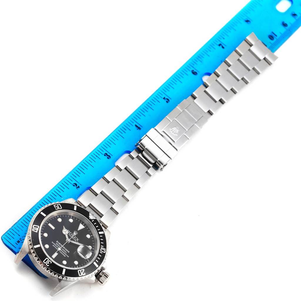 The Rolex Submariner 16610 Men's Stainless Steel Black Dial 16610 Men's Stainless Steel Black Dial watch is shown from a top view, highlighting its face, bezel, crown, and bracelet laid beside a blue ruler.