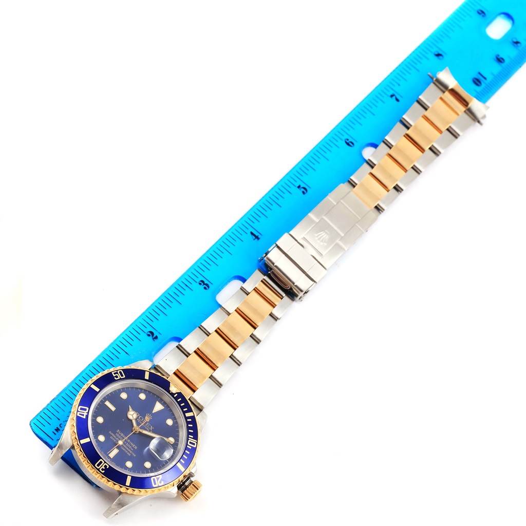 The image shows a top-down view of a Rolex Submariner 16613 Men's Steel and Gold (two tone) Blue Dial 16613 Men's Steel and Gold (two tone) Blue Dial, featuring its gold and silver bracelet alongside a ruler for scale.